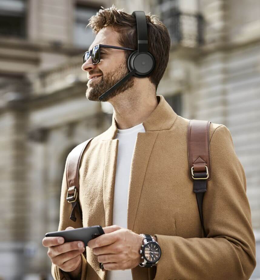 Foto di un uomo di profilo per strada mentre indossa delle cuffie wireless con microfono e con uno smartphone in mano.
